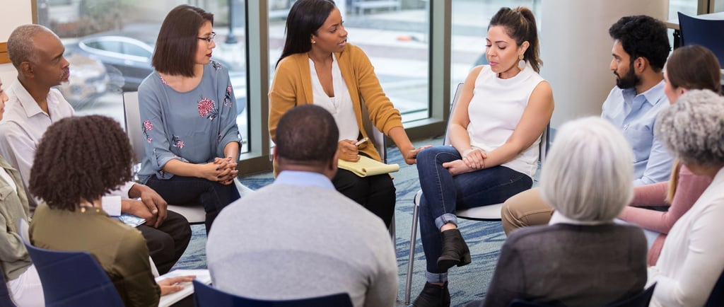 a group of people sitting in a circle with a woman in a white shirt