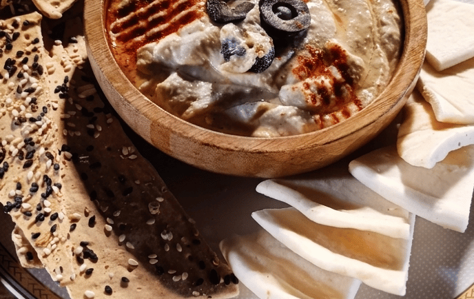 Wooden bowl of creamy hummus dip topped with olives and paprika, served with pita bread and crackers.