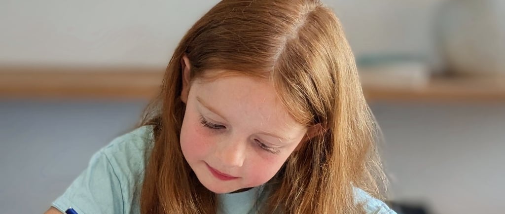A primary school girl with red hair focuses on writing her work for her key stage 2 tutoring at home