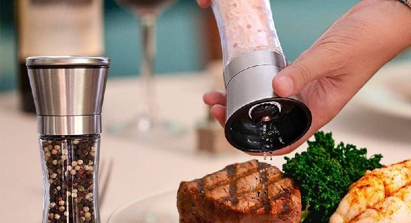 Hand using a stainless steel salt grinder over a gourmet steak and lobster dinner.