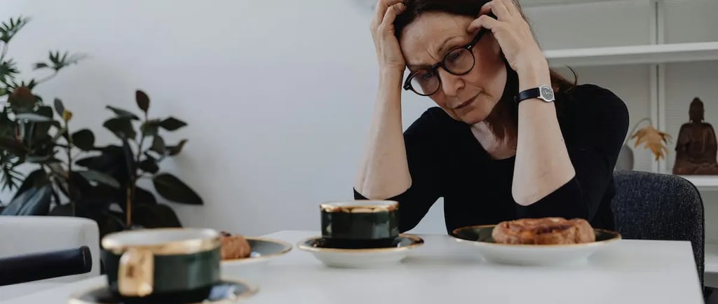 Stressed mature woman holding her head in frustration while sitting at a table with coffee and pastries.