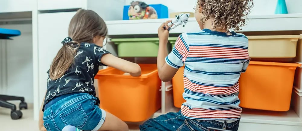 Two young children playing with toys and organized plastic storage bins in a bright playroom.