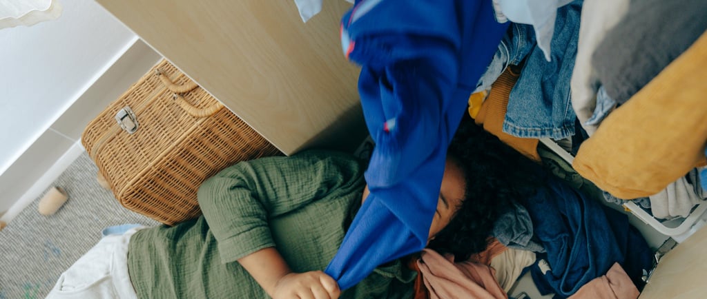 Child lying amongst pile of clothes in front of overflowing cupboard with a hamper basket