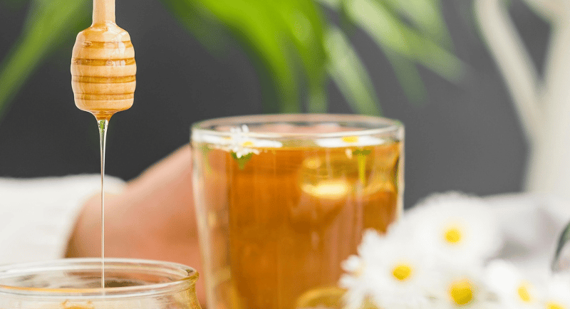 Golden organic honey dripping from a wooden dipper into a jar next to herbal chamomile tea.