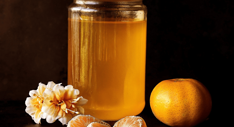 A glass jar of organic golden honey with fresh tangerine slices and flowers on a dark background.