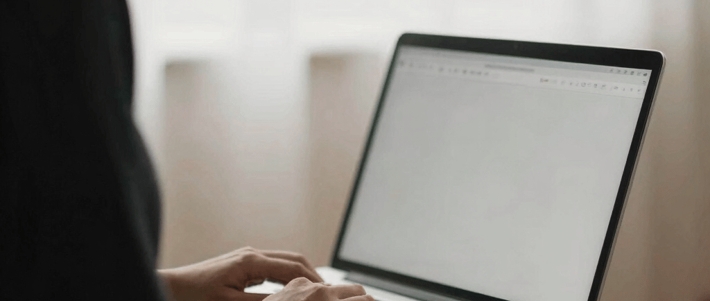 A person typing on a laptop computer while working from home near a bright window.