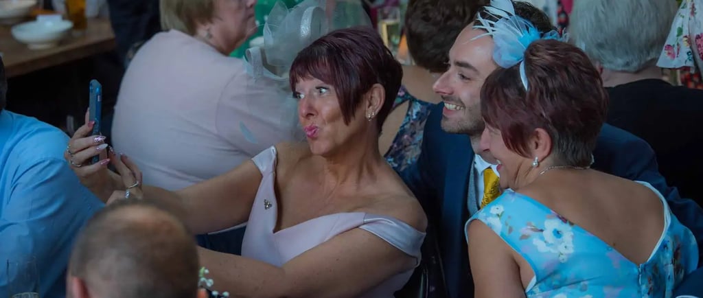 Wedding guests in elegant attire take a group selfie using a smartphone at a reception dinner.