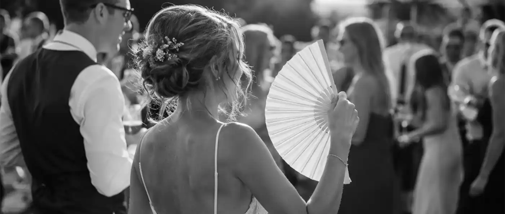 A bride in a lace wedding dress uses a handheld folding fan during an outdoor summer reception.