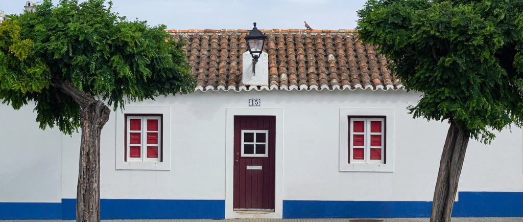Traditional white Portuguese house with blue trim, red windows, and symmetrical green trees.
