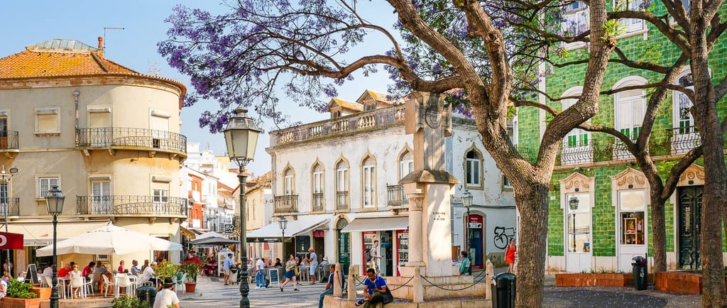 A sunny town square in Lagos, Portugal with blooming purple jacaranda trees and historic architecture.