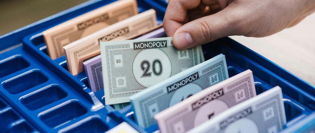 A hand selects a bill from a blue Monopoly game bank tray filled with colorful play money.