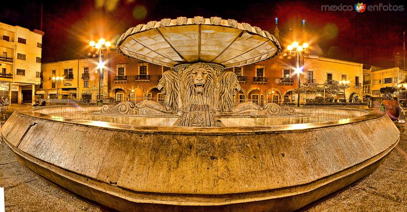 Illuminated lion fountain at night in a historic Mexican city plaza with glowing street lights.