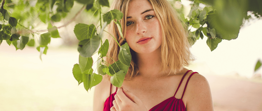 A blonde freckled woman in a red dress posing under green leaves in a park.
