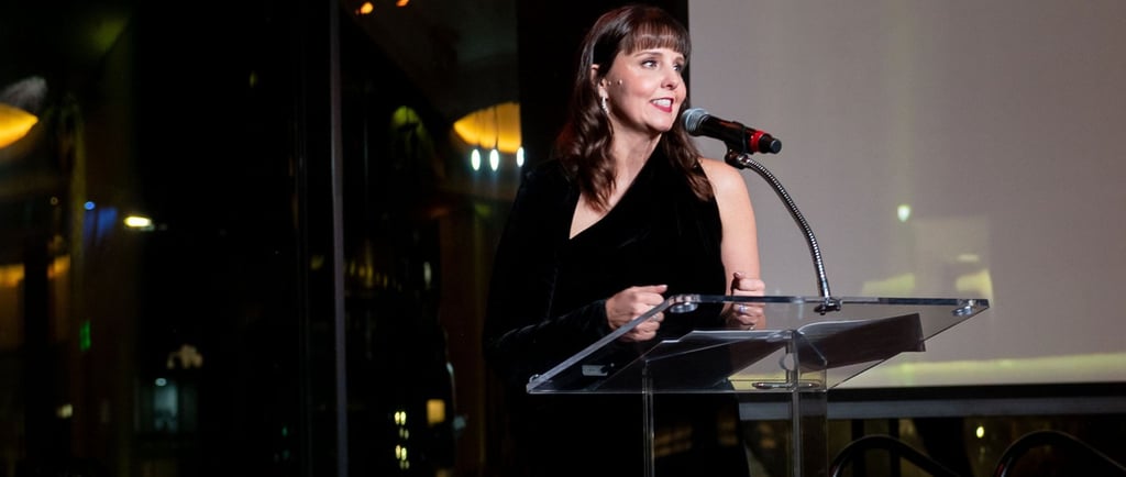Woman speaking at a podium with a microphone at an indoor evening event, city lights visible through windows behind her.