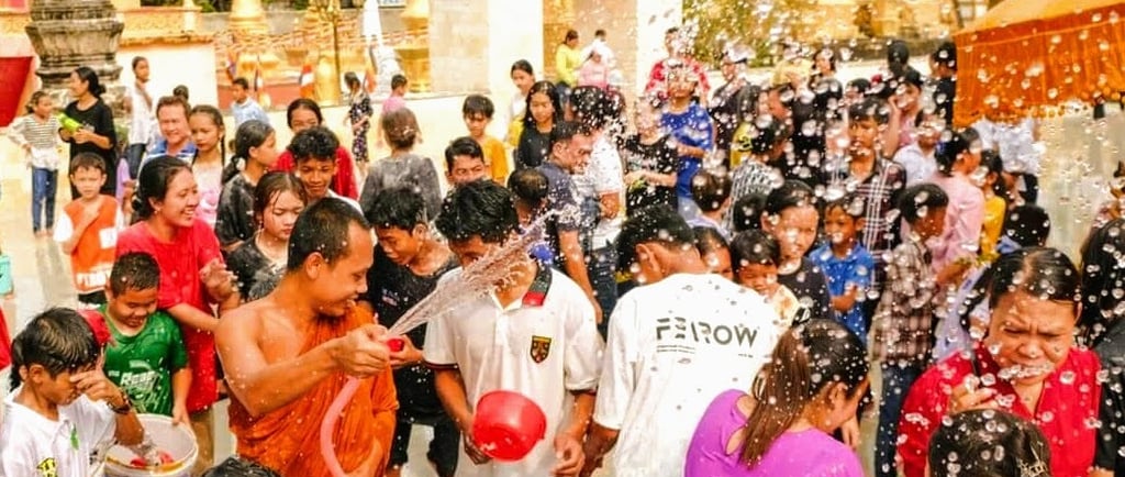 A monk in orange robes sprays a hose at a crowd of smiling people during a water fight at a Khmer pa
