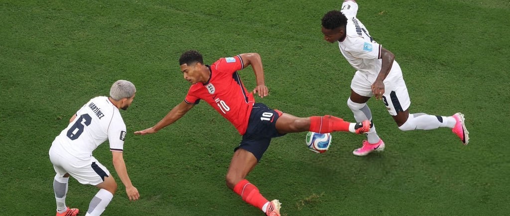 Jude Bellingham in an England kit dribbles past two defenders on a green soccer pitch during a match.