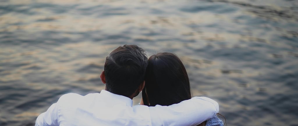 Couple sitting by the water with arms around each other during a romantic sunset date.