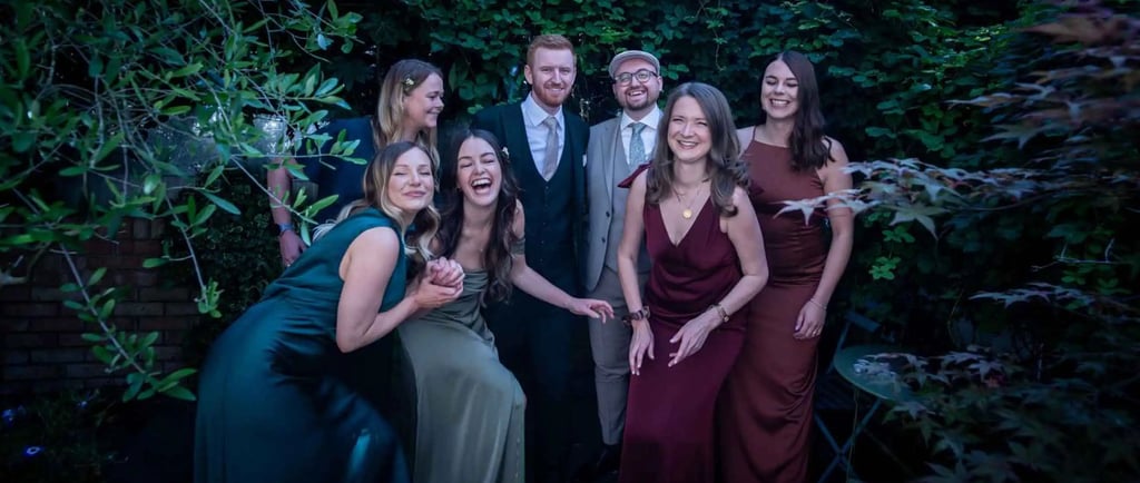 A group of friends in formal wedding attire laughing together in a lush green garden setting.