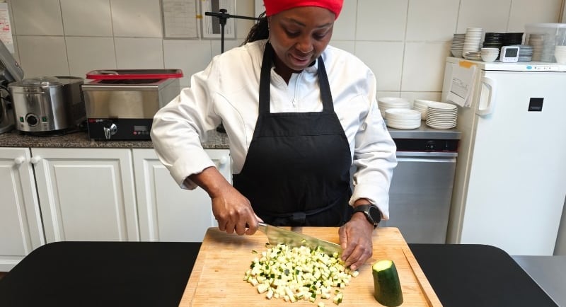 Chef Viviane in white chef jacket using a chef's knife to dice courgette on a bamboo chopping board on a dark kitchen surface