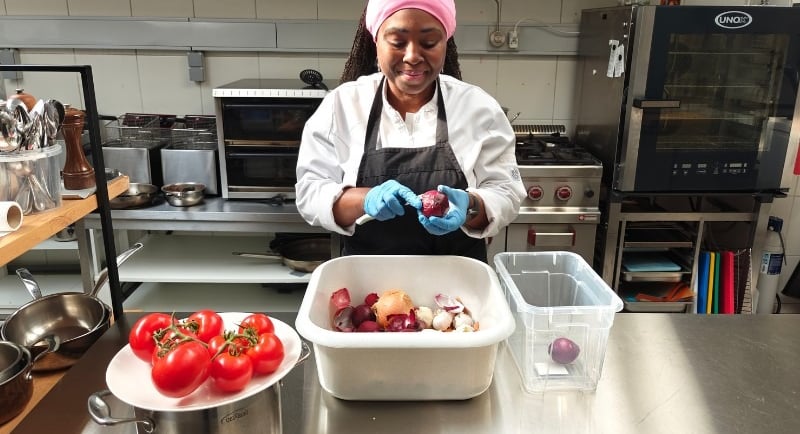 Chef Viviane in white chef jacket and black apron calmly peeling red onions beside fresh tomatoes on a stainless steel.