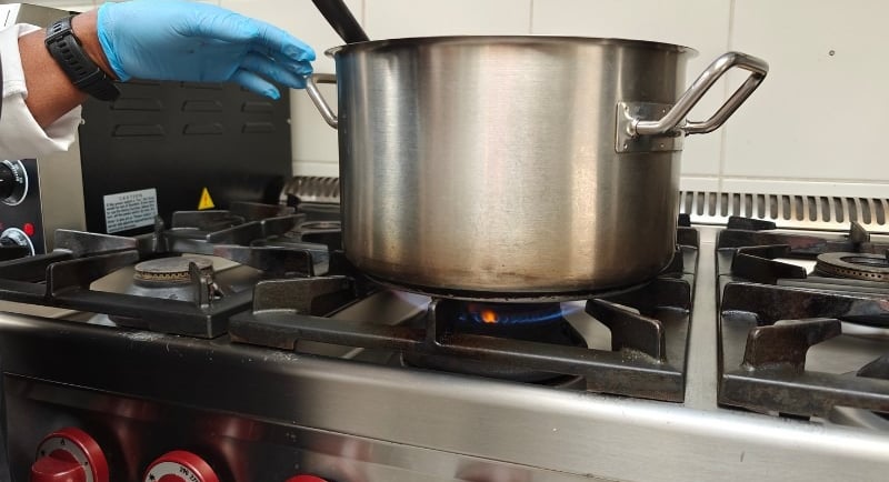 A chef's gloved hand stirring a large stainless steel stockpot over a blue gas flame on a professional kitchen range
