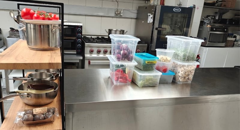 An organised professional kitchen station with pans on a wooden rack, fresh tomatoes and labelled prep containers.