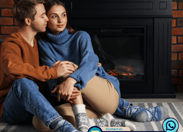 A couple in warm sweaters cuddling on a rug by a black fireplace with a brick background.