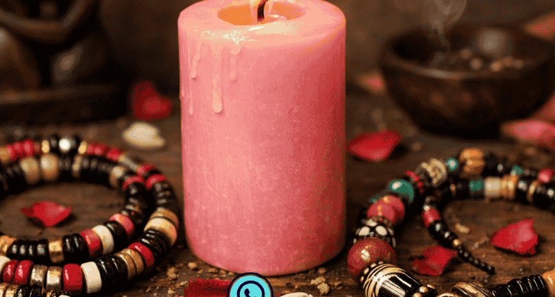 A glowing pink candle with African spiritual beads, rose petals, and a lovers figurine for ritual work.