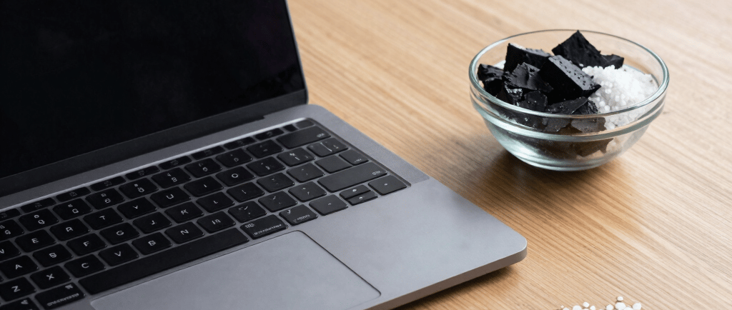 Laptop on a wooden desk next to a bowl of black charcoal and white sea salt.