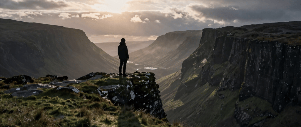 A hiker stands on a mossy cliff edge overlooking a dramatic mountain valley at sunset under cloudy skies.