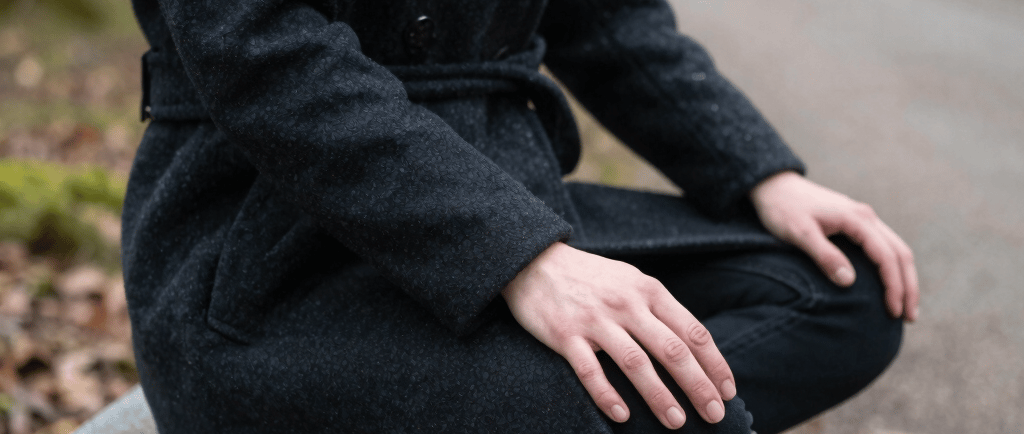 A person in a black wool coat sits in a cross-legged meditation pose on a stone park bench.