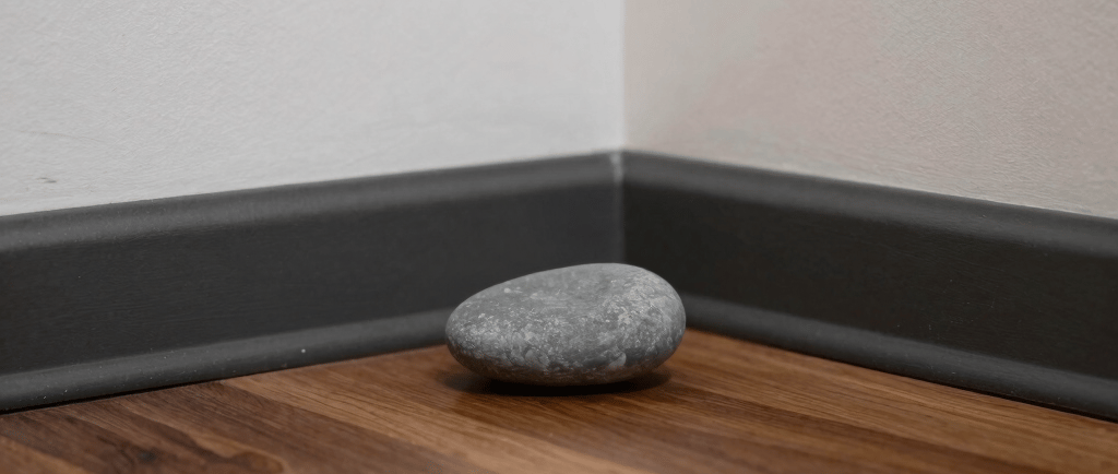 A smooth grey river stone sits on a walnut laminate floor in the corner of a room with white walls.