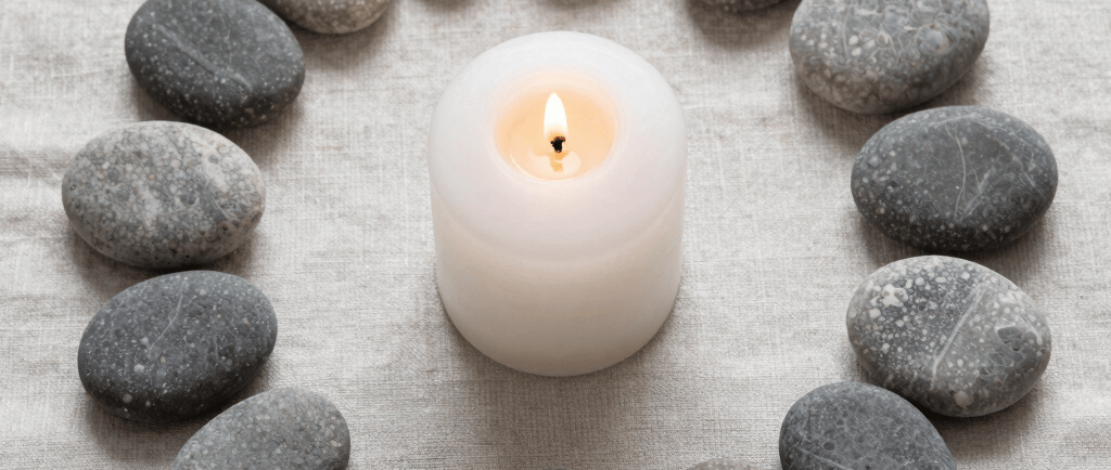 A lit white pillar candle centered within a circle of smooth grey zen stones on beige linen.