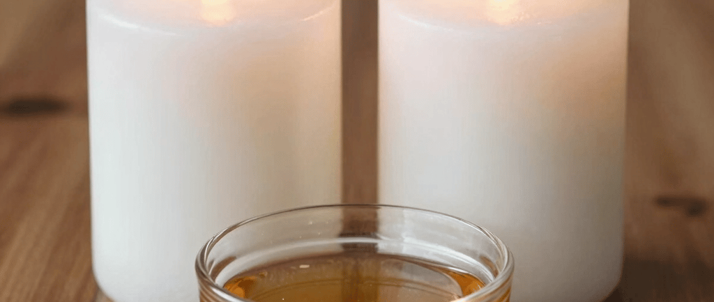 Two lit white pillar candles behind a small glass bowl of golden massage oil on a wooden table.