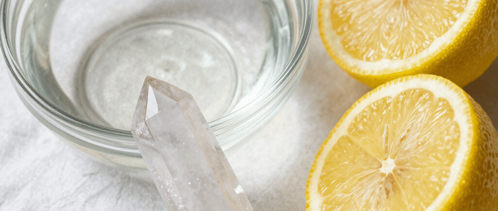 Clear quartz crystal point next to a glass bowl of water and two lemon halves on a white cloth.