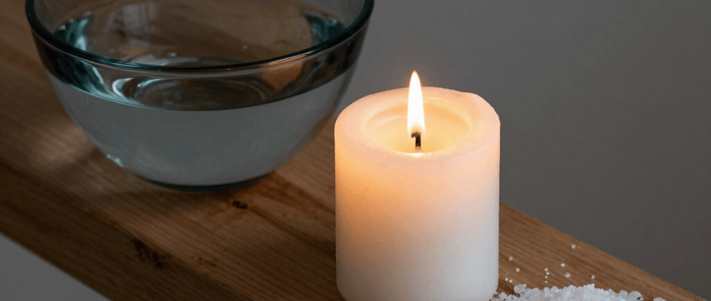 A lit white pillar candle beside a bowl of water and a pile of bath salt on a wooden spa tray.