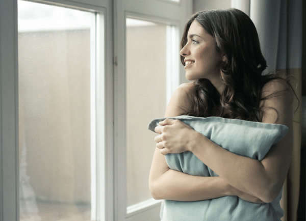 a girl sitting beside window and holding pillow