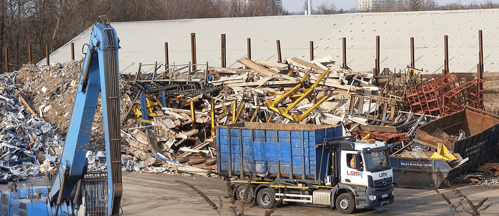A scrap metal recycling yard with piles of mixed metal, wood, and industrial debris. A blue hydrauli