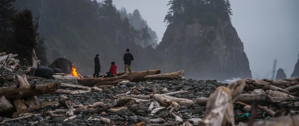 a group of people standing around a campfire in the PNW among big rocks and the ocean