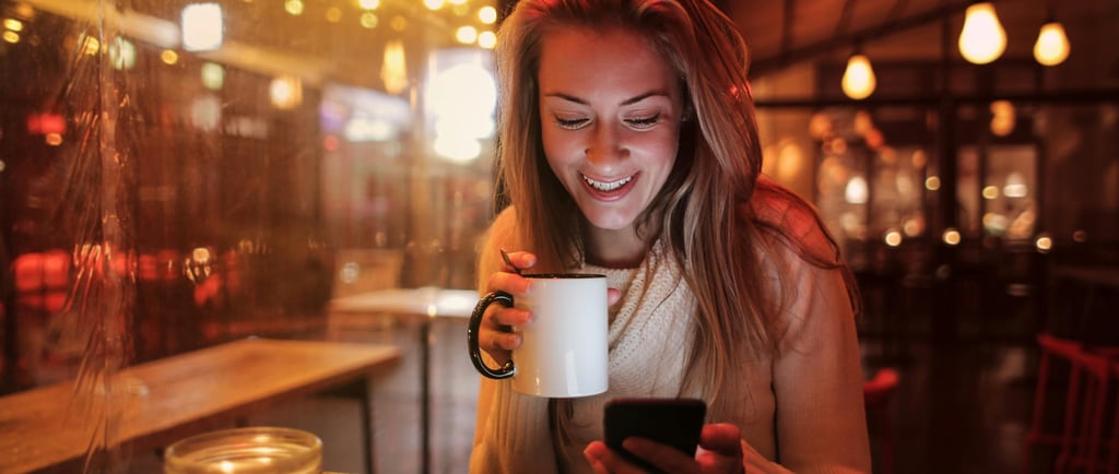Smiling woman holding a coffee mug while looking at her smartphone in a cozy, warmly lit café at night.