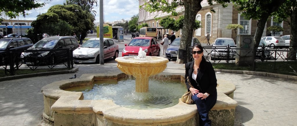 Foto da autora sentada numa fonte na praça próxima ao porto de Cádiz.