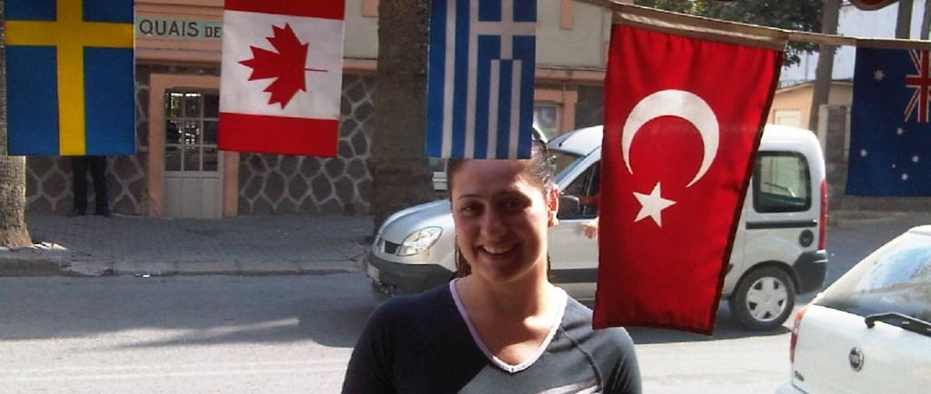 Foto da autora no centro de Izmir, entre uma bandeira turca e a movimentação local.