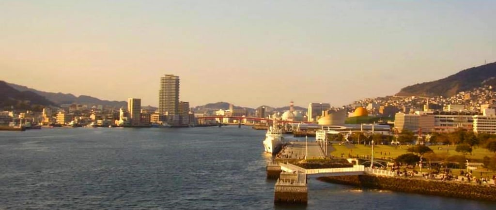 Navio de cruzeiro chegando ao porto de Nagasaki, com pessoas aguardando na orla e colinas verdes ao fundo