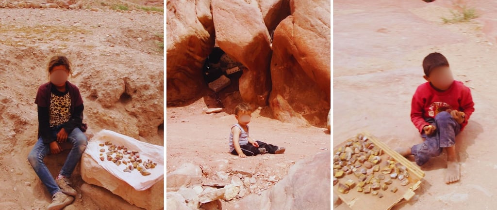 Três crianças em Petra, oferecendo pedrinhas coloridas aos turistas, em meio ao cenário rochoso.