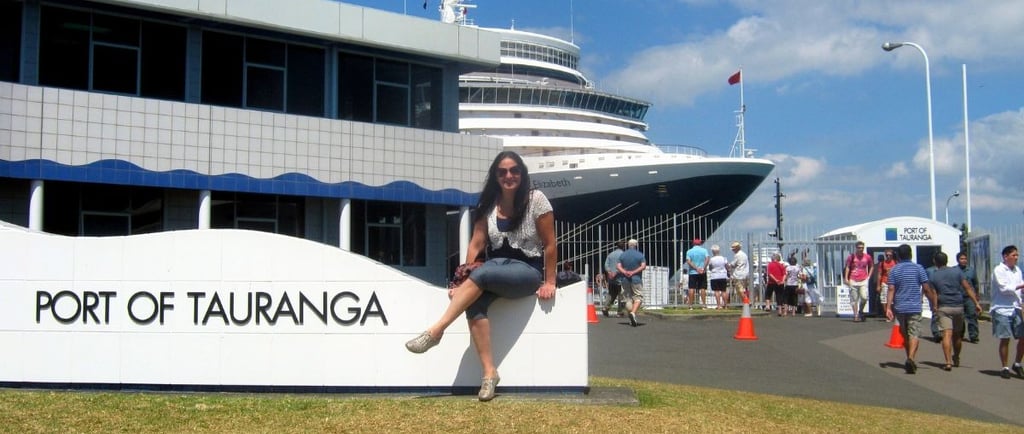 Foto da autora sentada no Porto de Tauranga, com um navio e a cidade ao fundo.
