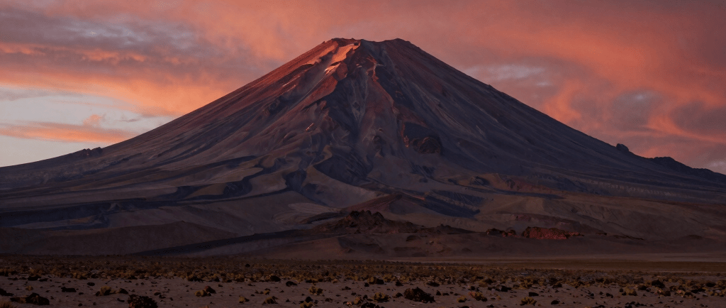 A majestic volcanic mountain peak under a vibrant pink and orange sunset sky in a desert landscape.