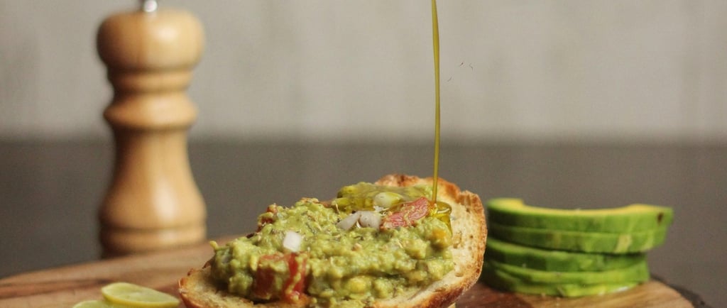 Fresh avocado toast on rustic wood being drizzled with olive oil next to a pepper grinder and lime slices.