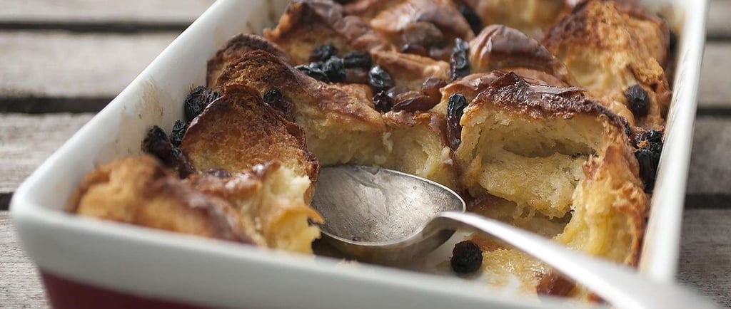 Golden brown bread and butter pudding with raisins in a ceramic baking dish with a serving spoon.
