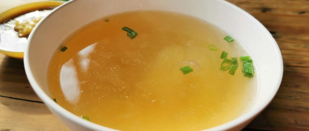 A white ceramic bowl of clear chicken broth garnished with chopped green onions on a rustic wooden table.