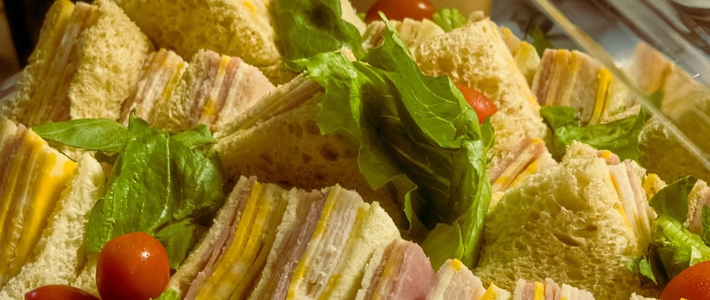 Freshly prepared ham and cheese finger sandwiches with lettuce and cherry tomatoes on a catering platter.